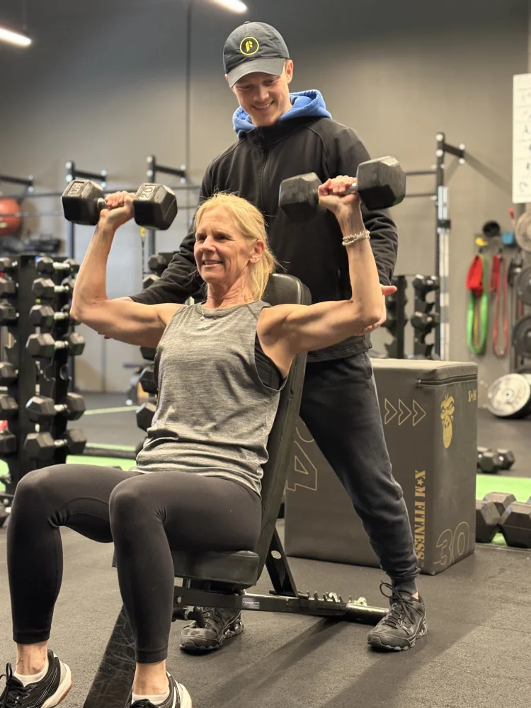 Personal Training Free Weights Woman performs dumbbell shoulder press while male Fit Coach supports her